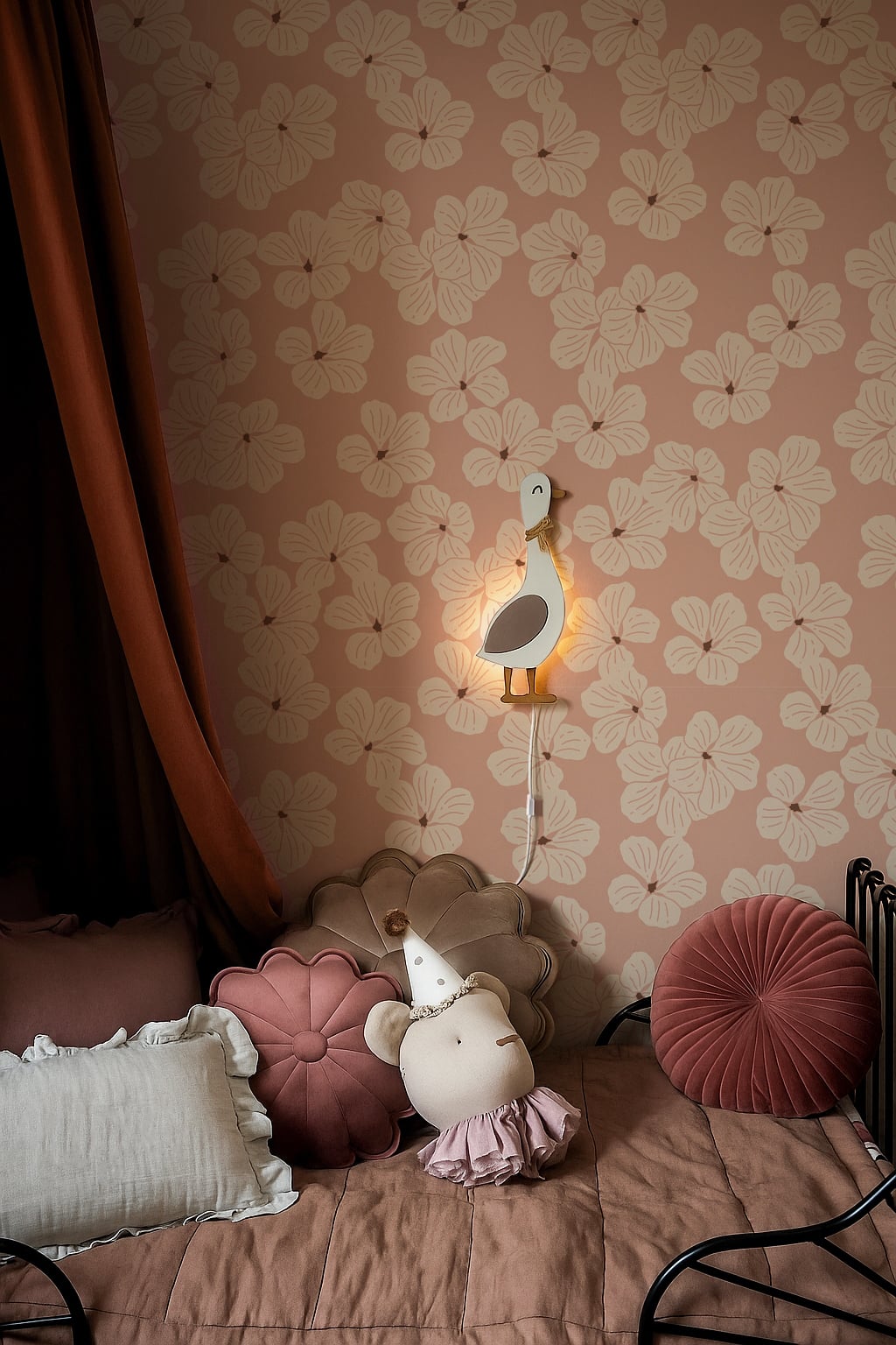 Oudroze bloemenbehang in een meisjeskamer met een wandlampje en zachte kussens