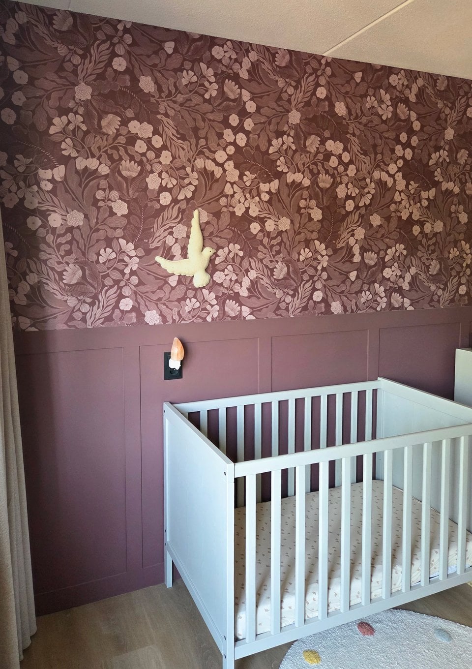 Mauve bloemenbehang boven een lambrisering in de babykamer